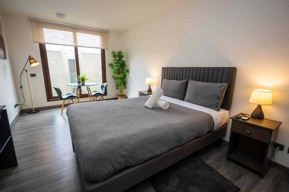 Quarto de hotel moderno com uma cama grande, abajures, uma pequena mesa com cadeiras, uma planta em vaso e vista pela janela.