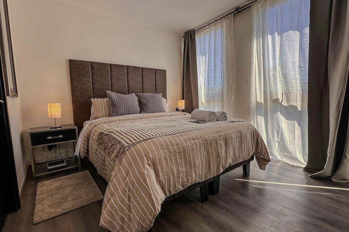 Uma cama bem arrumada com colcha listrada, uma mesa de cabeceira com abajur e vista através de cortinas translúcidas.