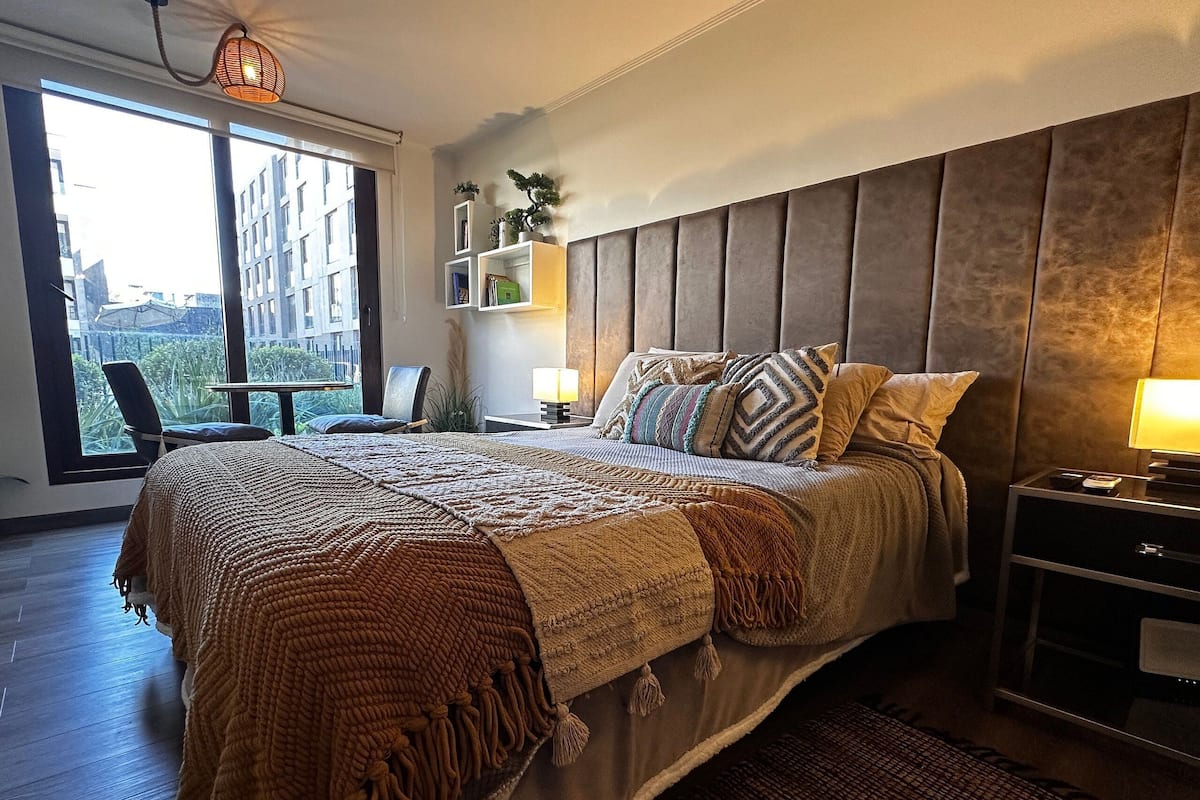 Quarto moderno com cama grande, mesa de cabeceira, cadeira e vista para edifícios por uma grande janela.