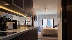 Una cuina moderna amb una illa central, una zona de menjador amb taula i cadires, i un dormitori amb llit i finestra.