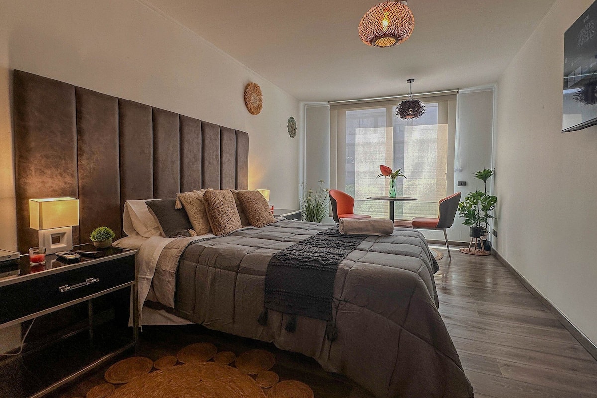 Um quarto moderno com uma cama grande, mesas de cabeceira, uma pequena mesa com cadeiras e uma janela ampla.