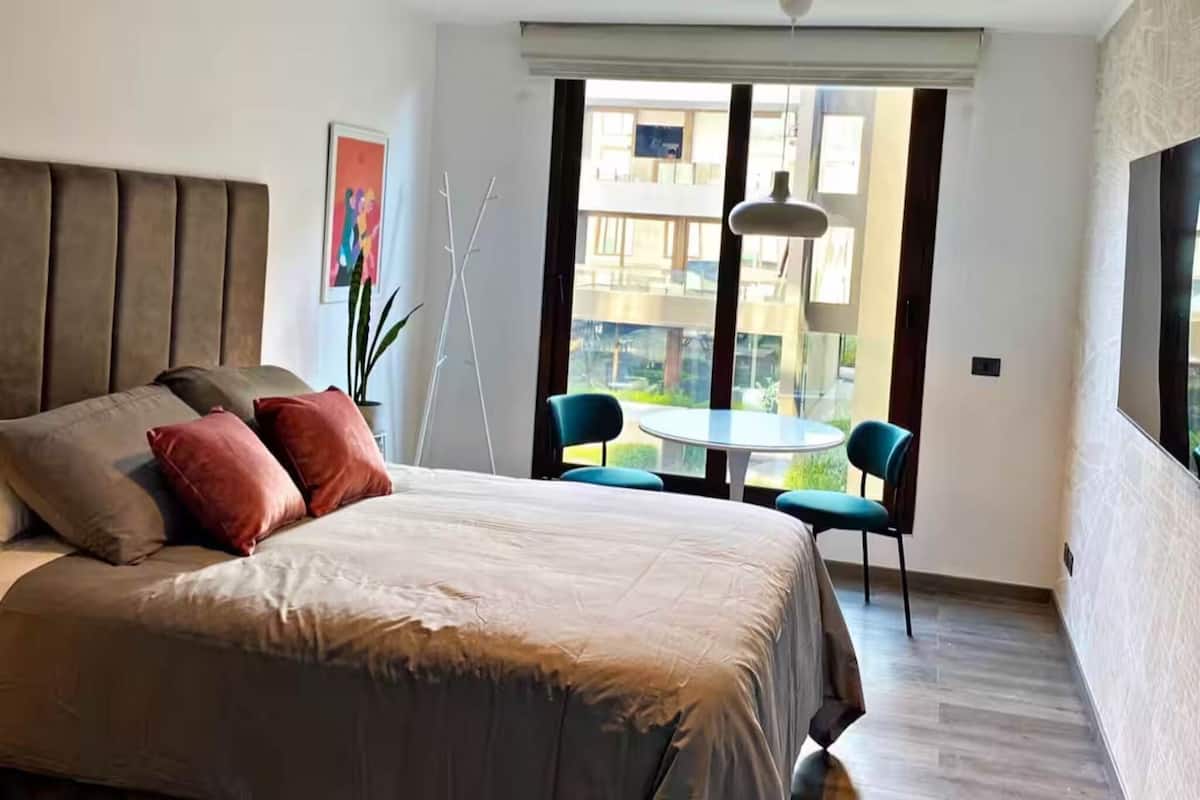 Quarto moderno com uma cama grande, uma mesa pequena, duas cadeiras e vista para o exterior.
