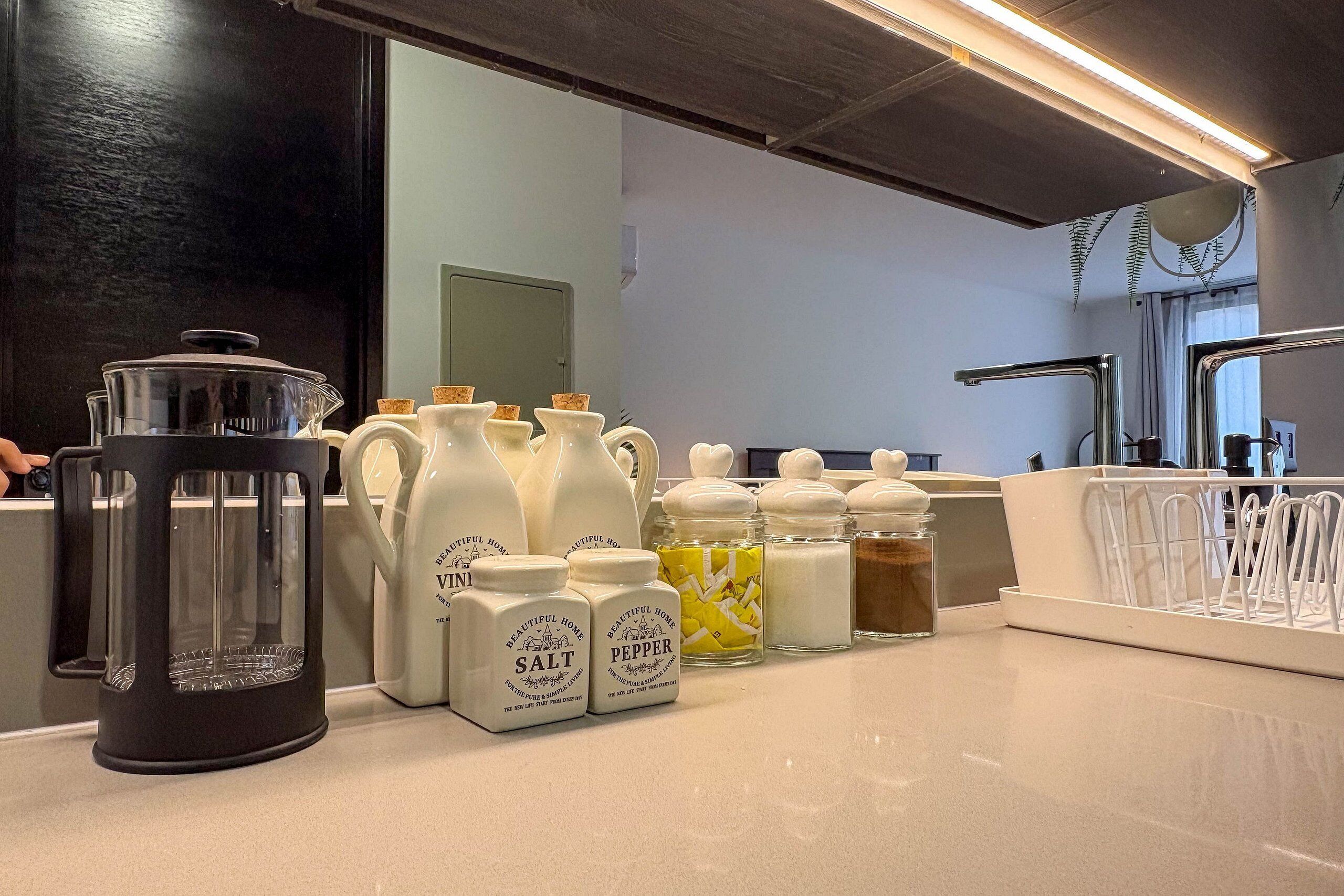 Uma bancada de cozinha com diversos eletrodomésticos e recipientes, incluindo uma cafeteira, saleiro e pimenteiro e um processador de alimentos.
