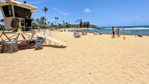 Ein Strand mit einem Rettungsturm, Palmen und Menschen, die in der Sonne entspannen.