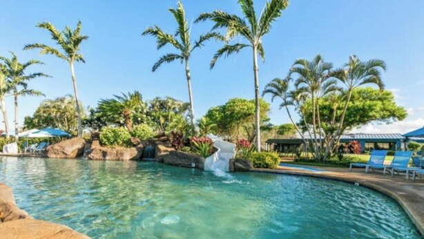Ein klares Schwimmbecken mit Wasserfall, umgeben von Palmen und Liegestühlen.