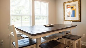 A dining area with a wooden table and white chairs, a framed artwork on the wall, and a window with blinds.