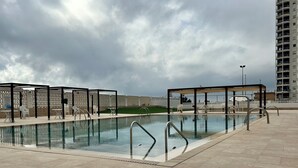 Ein Pool mit Fliesenplatten umgeben, umrahmt von überdachten Sitzbereichen und einem hohen Gebäude im Hintergrund.