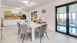 Een moderne keuken met witte kasten, een eethoek met een witte tafel en stoelen, en een grote schuifpui naar een zwembad.