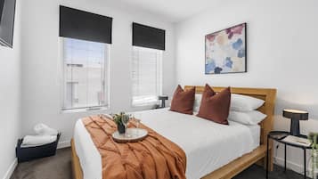 A modern bedroom with a wooden bed, white bedding, and a brown throw blanket. There are two windows with blinds, a bedside table with a lamp, and a small plant on the nightstand.