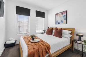 Une chambre moderne avec un lit en bois, une literie blanche et un plaid marron. Il y a deux fenêtres équipées de stores, une table de chevet avec une lampe et une petite plante posée sur la table de chevet.
