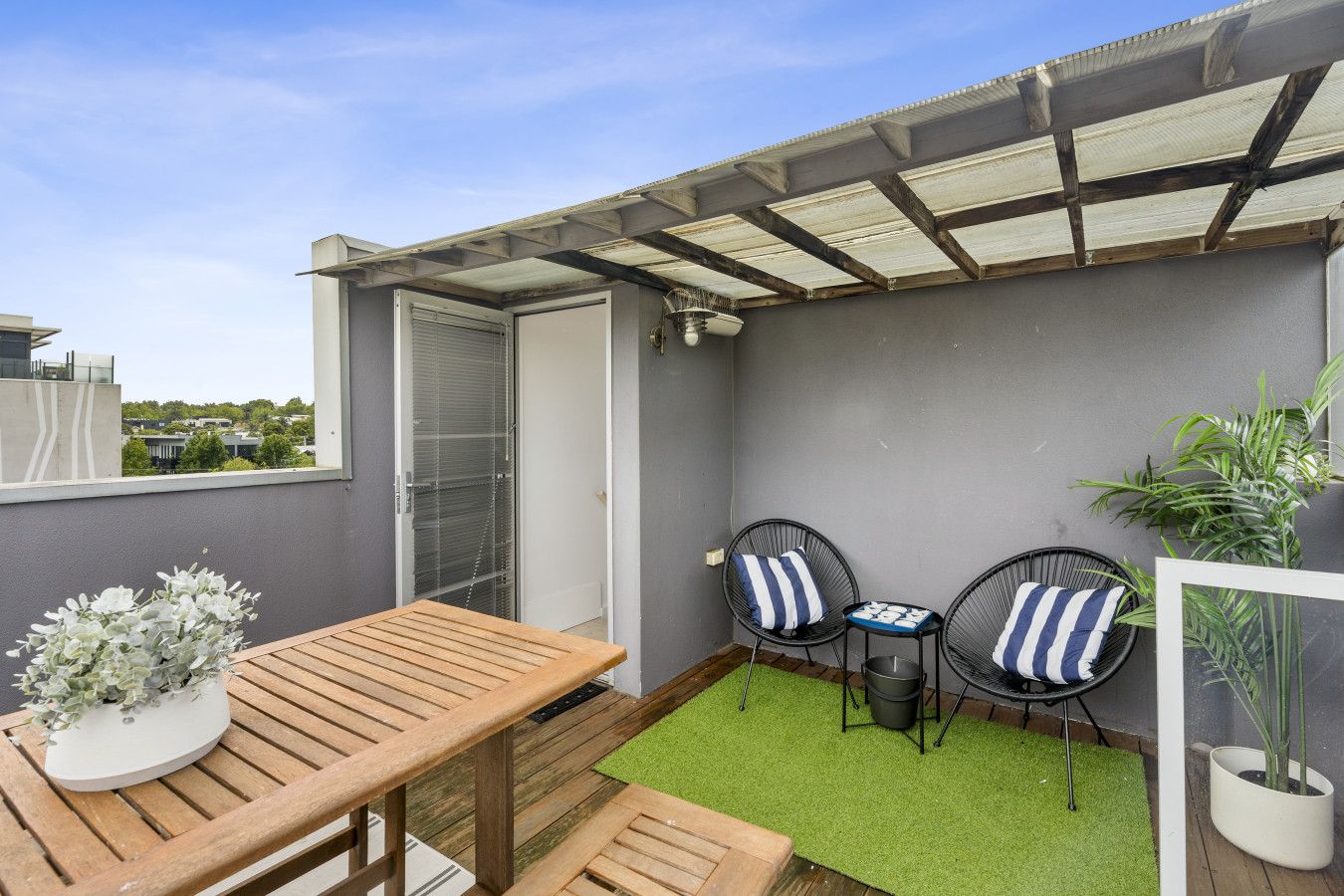 A patio area with a wooden table and two chairs, a small green rug, and a potted plant.