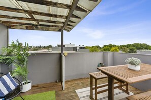 A rooftop patio with a wooden table and chairs, a potted plant, and a view of the cityscape. - The Red Ember Retreat (Caulfield North)