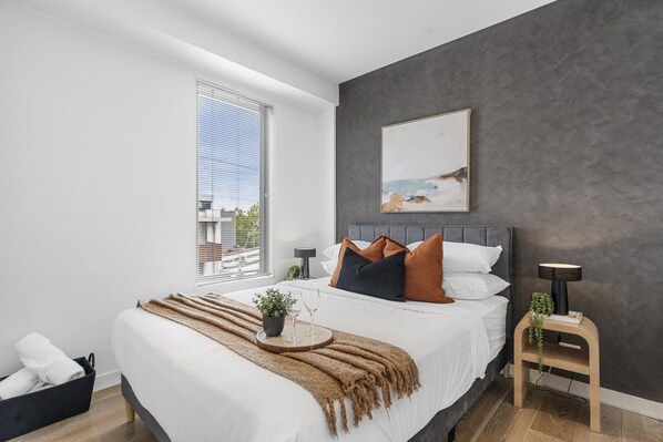 A modern bedroom with a large bed, a window with blinds, a bedside table with a lamp, and a painting on the wall.