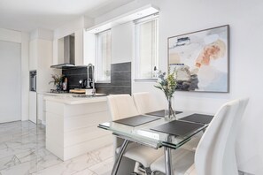 A modern kitchen with white cabinetry, a central island, and a dining area with a glass table and white chairs. - The Red Ember Retreat (Caulfield North)