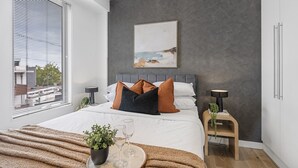 A modern bedroom with a large bed, a bedside table with a lamp, a small side table with a plant, and a window with blinds.