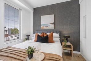 A modern bedroom with a large bed, a bedside table with a lamp, a small side table with a plant, and a window with blinds. - The Red Ember Retreat (Caulfield North)