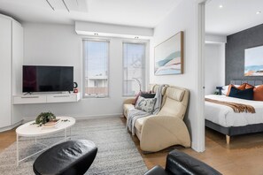 A modern living room with a sofa, a coffee table, and a TV. There are two windows with blinds, a painting on the wall, and a bed in the background.