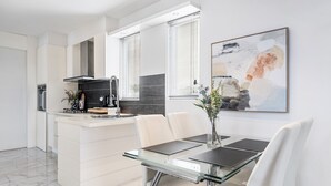 A modern kitchen with white cabinetry, a central island, and a dining area with a glass table and white chairs. - The Red Ember Retreat (Caulfield North)