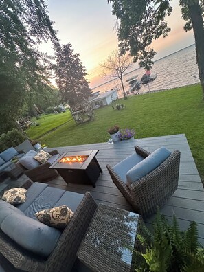 A deck with outdoor furniture and a fire pit, overlooking a body of water and a dock with boats.