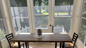 Un comedor con mesa de madera y bancos, ventanas amplias y vistas a una valla blanca y follaje.