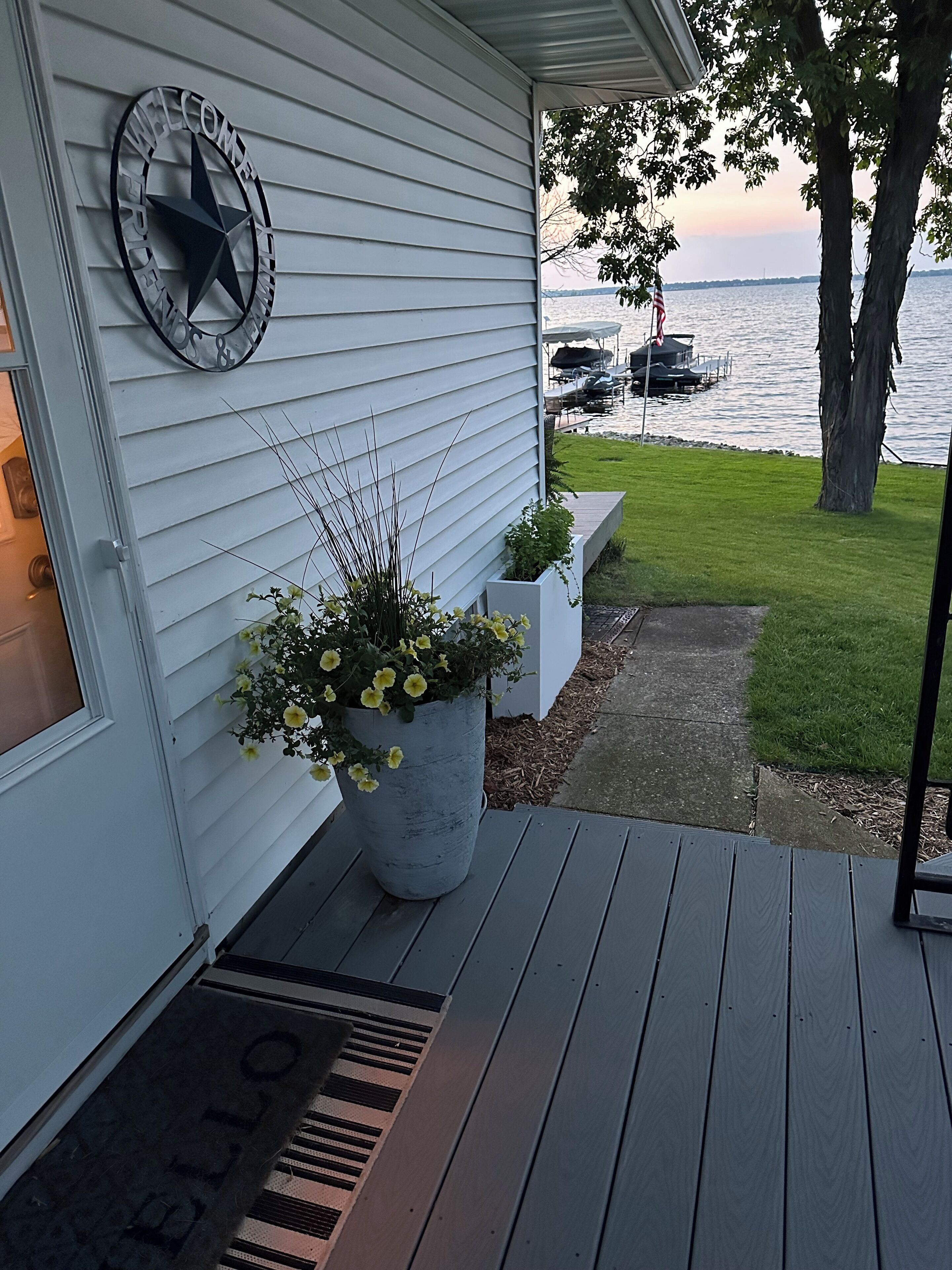 A house with a welcome sign, a large planter with yellow flowers, a wooden deck, and a view of a lake with boats.