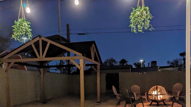 En udendørs patio med bålsted, siddepladser, hængende planter og en pergola.