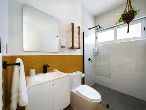 A modern bathroom with a white sink, toilet, and mirror. There is a towel rack with towels, a soap dispenser, and a hanging plant. - SAYU - Suites (Cozumel)