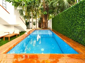 A rectangular swimming pool with a wooden deck surrounding it, a white wall on one side, and a green hedge on the other. - SAYU - Suites (Cozumel)