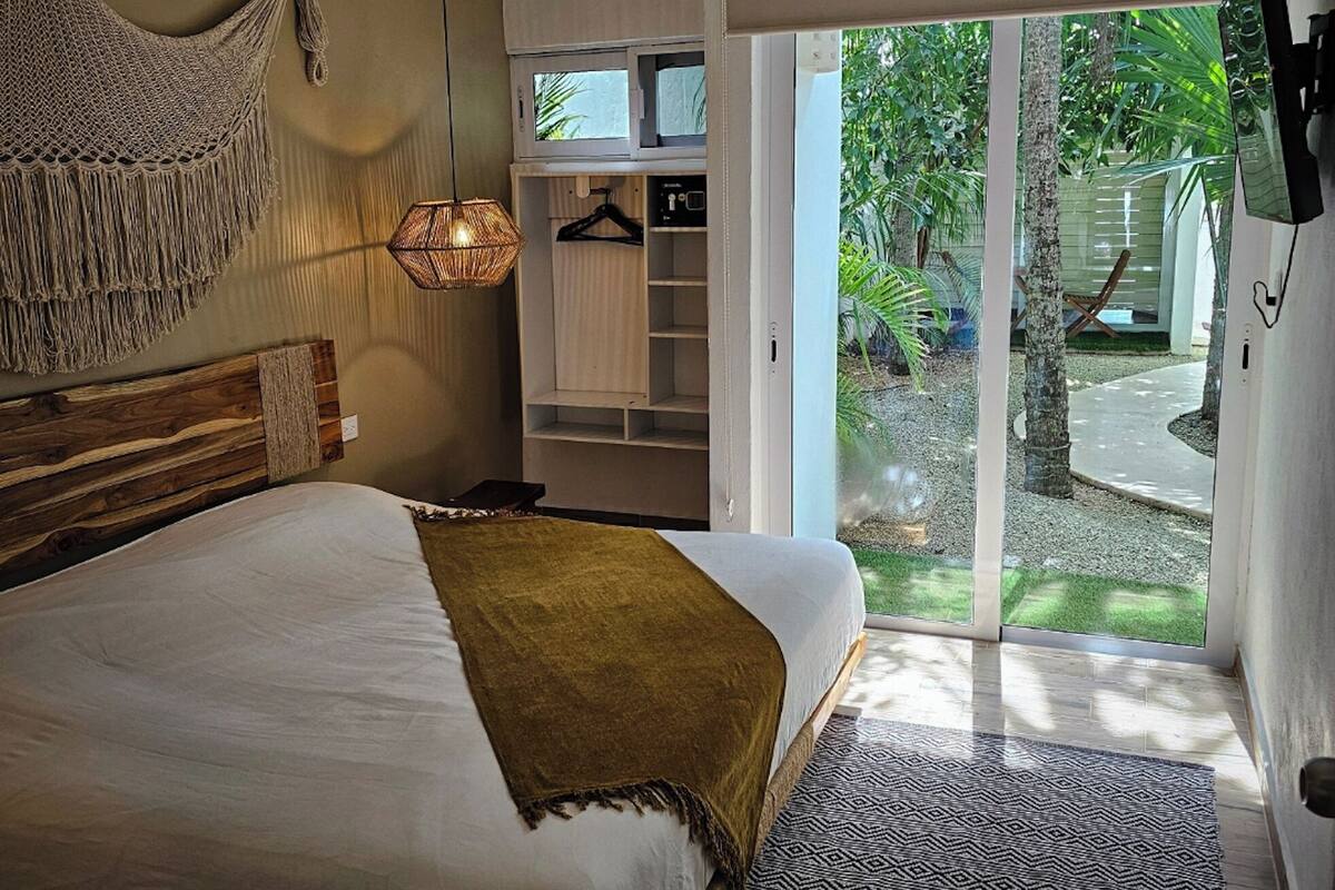 A bedroom with a bed, a ceiling fan, a woven wall hanging, a bedside lamp, and a view of a garden through a sliding glass door.