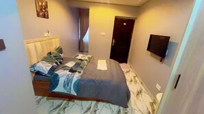 A small, narrow hotel room with a bed, a flat-screen TV mounted on the wall, and a door leading to another room. - A four bedroom smart  Gated Apartment , Wi-fi, CCTV short drive from the Airport (Accra)
