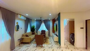 Una sala de estar con un sofá, sillones, una mesa de centro y un televisor. Hay un ventilador de techo, una planta y una ventana con cortinas.