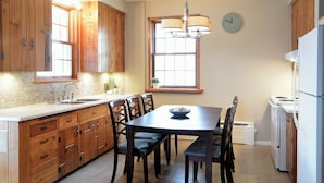 Una cocina con armarios de madera, una mesa de comedor con sillas, una araña, un reloj y un refrigerador.