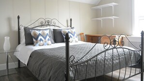 Una cama bien hecha con estructura metálica, una mesita de noche con un jarrón blanco y una ventana con estantes.