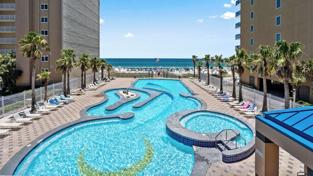 A swimming pool with a wavy design, surrounded by palm trees and lounge chairs, with a beach and ocean view in the background. - Lazy River, 3 Pools - Crystal Tower - Free Daily Activities & Beach Service! (Gulf Shores)