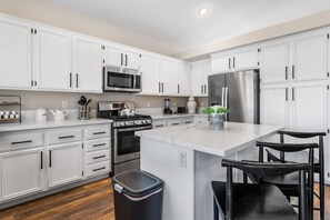 A modern kitchen with white cabinetry, stainless steel appliances, a central island with bar stools, and a trash can.