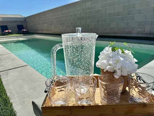 A clear glass pitcher, a glass carafe, and a floral arrangement on a wooden tray by a swimming pool.