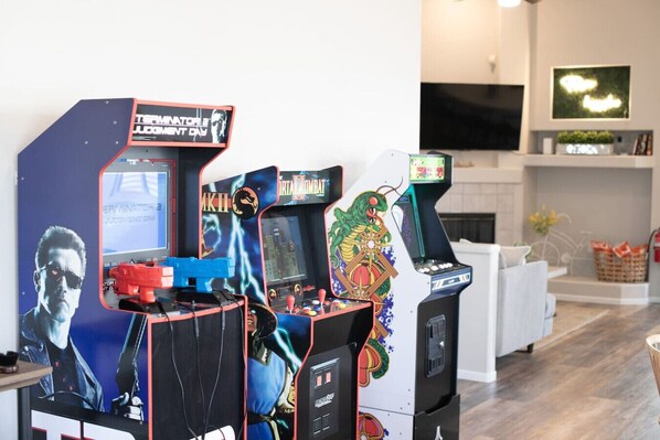 Three arcade machines in a modern living room with a fireplace and a TV mounted on the wall.