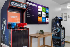 A room with a large TV displaying streaming services, a gaming arcade machine, and a small table with a plant.