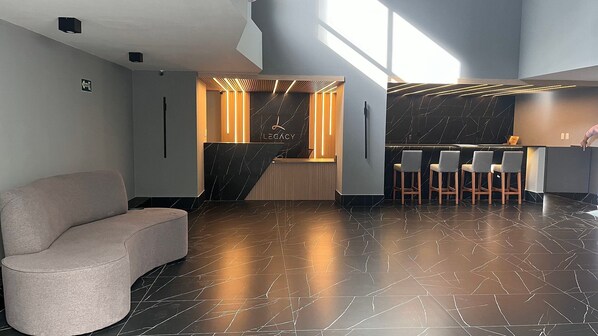 A modern reception area with a marble reception desk, a seating area with bar stools, and a large window providing natural light. - Flat Gold Legacy Suites - Cabo Branco (João Pessoa)