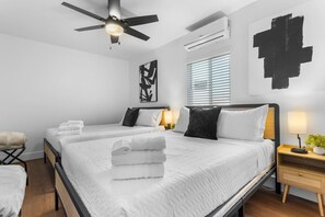 Een hotelkamer met twee bedden, een plafondventilator, een airconditioningapparaat, een nachtkastje met een lamp en een wanddecoratie.