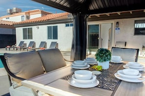 Un área de comedor al aire libre con una mesa dispuesta para cuatro personas, rodeada de sillones, una piscina y un edificio con techo de tejas.
