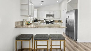 A modern kitchen with white cabinets, a granite countertop, a stainless steel refrigerator, and three bar stools. - 4BR Getaway w/Pool and Billiard! (North Las Vegas)