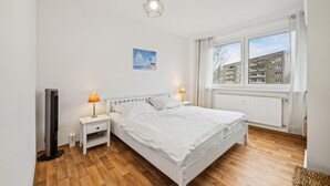 A bedroom with a bed, two bedside tables with lamps, a radiator, a window with a view of buildings, and a wall-mounted picture. - 1 bedroom cozy apartment in Anklam (Anklam)