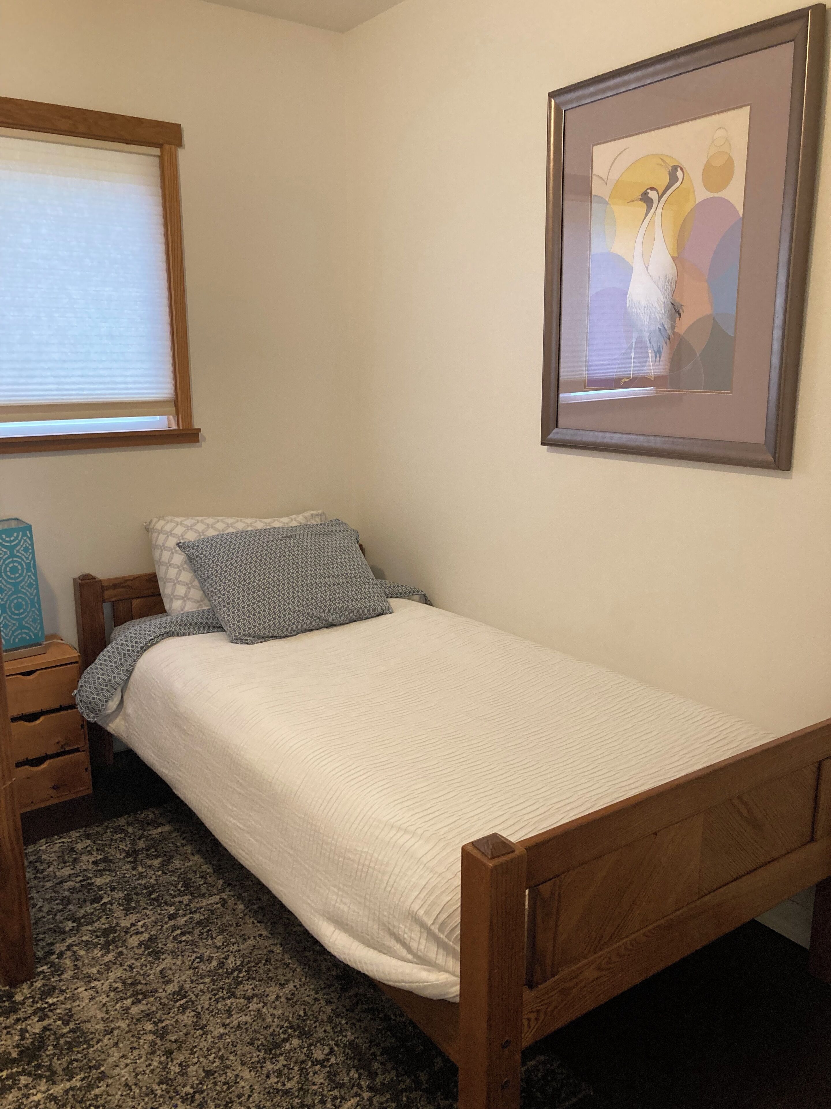 A small, neatly made bed with a wooden headboard, a bedside table with drawers, a framed picture on the wall, and a window with blinds.