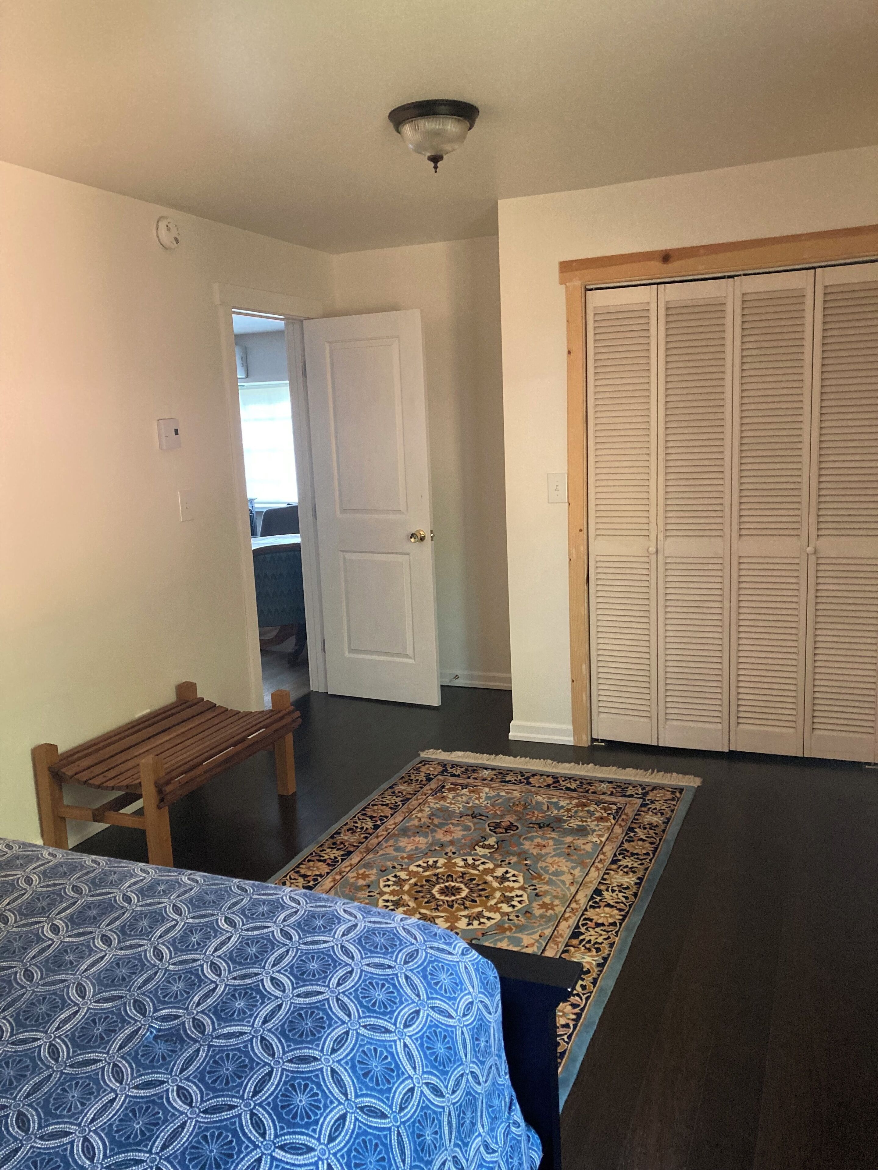 Een slaapkamer met een bed, een houten bankje, een vloerkleed met een patroon, een kast en een deur naar een andere kamer.