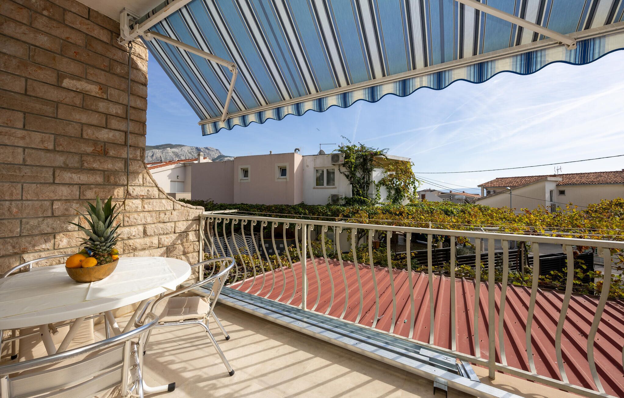 A balcony with a table and chairs, a metal railing, and a view of neighboring buildings and vegetation.