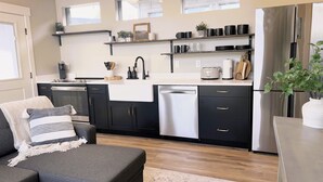 A modern kitchen with black cabinets, a stainless steel refrigerator, and a built-in dishwasher. There is a gray sofa with a throw blanket and pillows, and a small potted plant on the right side.