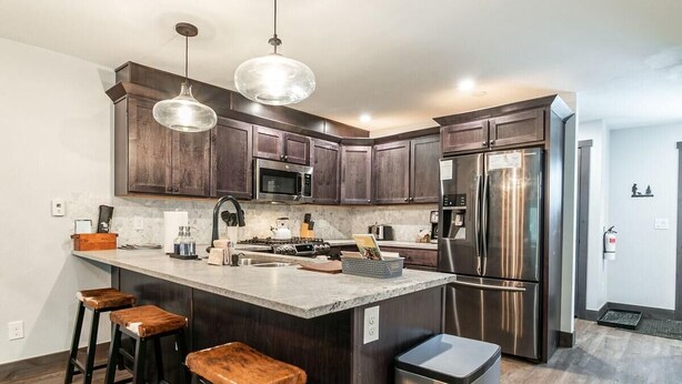 A kitchen with dark wood cabinets, stainless steel appliances, a central island with stools, pendant lighting, and a trash bin.