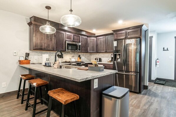 A kitchen with dark wood cabinets, stainless steel appliances, a central island with stools, pendant lighting, and a trash bin.