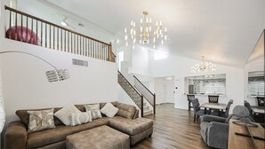 Ein geräumiges Wohnzimmer mit einem braunen Ecksofa, einem Hocker und grauen Sessel. Eine Treppe führt zu einer zweiten Etage mit einem roten Fitnessball. Der Raum verfügt über eine Chandelüster und einen Essbereich mit Tisch und Stühlen.